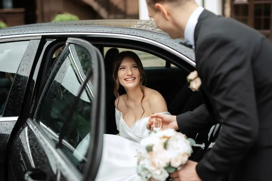 A groom in a suit holds the hand of his elegantly attractive bride, helping her out of a wedding getaway car, while he is holding a bouquet of roses in his other hand.